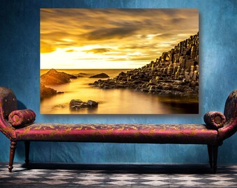 Op de afbeelding: Een ingelijste landschapsafdruk van een kustlandschap met een gouden zonsondergang. De afbeelding wordt weergegeven boven een rode chaise longue met sierlijke details. De scène toont rotsformaties en water.