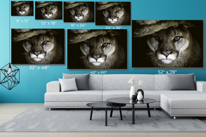 May include: A close-up photograph of a mountain lion's face. The animal's fur is a light brown color and its eyes are a bright green. The background is dark and blurry.