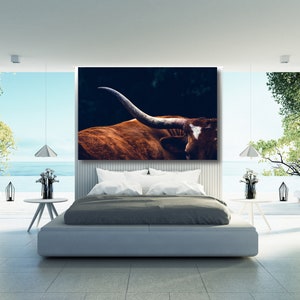 May include: A close-up of a brown and white longhorn steer with large horns. The steer is in a dark setting with a blurred background. The image is framed and hanging above a bed in a modern bedroom.