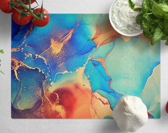 May include: A blue, green, and orange abstract marble cutting board with gold accents. The board is on a white surface with a white bowl of dip, a head of garlic, and tomatoes.