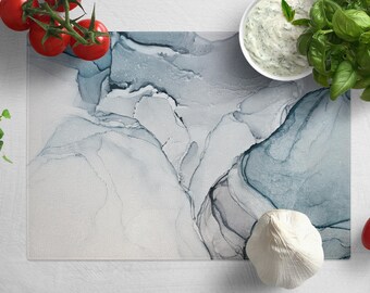 May include: A blue and gray marble-patterned cutting board with a white garlic clove, a bowl of white sauce, a sprig of green basil, and red tomatoes on a white surface.