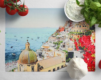 May include: A watercolor painting of a colorful Italian village with a domed church, overlooking a blue ocean with boats. The scene is framed by red flowers and green plants.