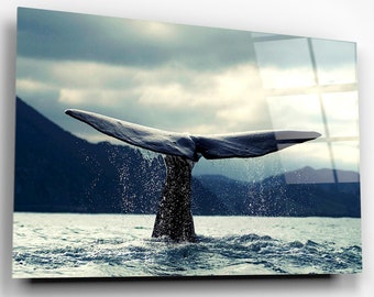 May include: A whale's tail breaks the surface of the water, creating a spray of droplets. The tail is dark gray and the water is a deep blue. The background is a hazy, blue sky with mountains in the distance.