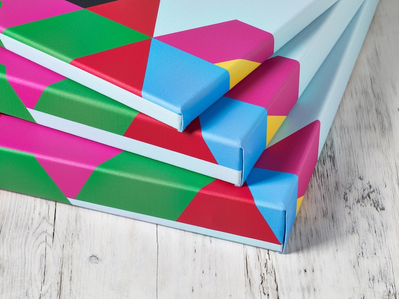 May include: Three colorful canvas prints with geometric patterns in shades of red, pink, blue, green, and yellow. The prints are stacked on top of each other, with the top print slightly offset from the bottom two.