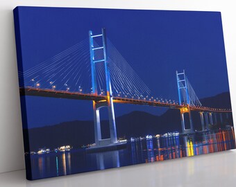 Op de afbeelding: Een grote hangbrug verlicht 's nachts met felle lichten. De brug overspant een waterlichaam met een donkerblauwe lucht op de achtergrond.