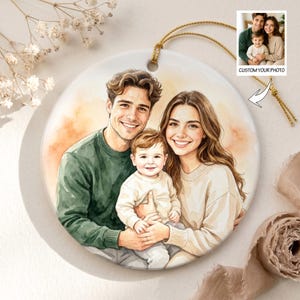 May include: A circular ceramic ornament with a watercolour portrait of a family of three. The ornament has a gold hanging string and a small inset photo with the text "CUSTOM YOUR PHOTO". The family is smiling, with a warm-toned background.