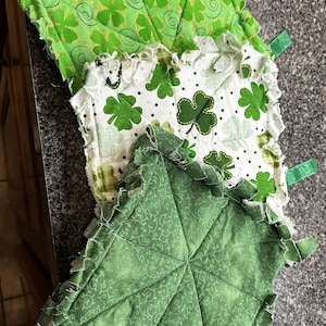 May include: Three green fabric squares with shamrock designs. The squares are layered on top of each other, with the largest square on the bottom and the smallest square on top. The squares are all made of a soft, plush fabric and have a frayed edge.