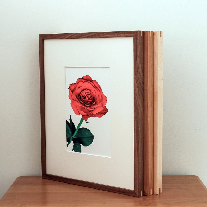 May include: A framed print of a single red rose with green leaves. The frame is made of wood with a dark brown finish. The frame is triple-layered with a lighter wood on the outer edges.