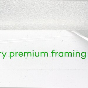 May include: A close-up of a white surface with the text "Gallery premium framing" printed in green. A clear acrylic sheet is being peeled off the surface.