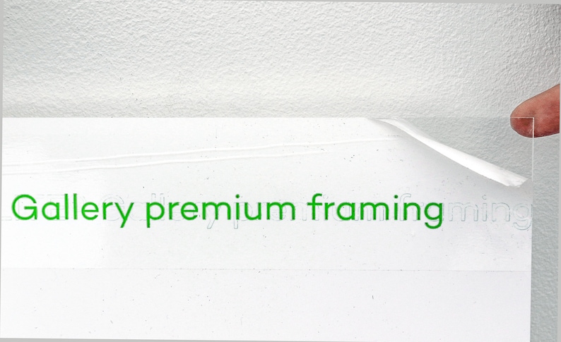 May include: A close-up of a white surface with the text "Gallery premium framing" printed in green. A clear acrylic sheet is being peeled off the surface.