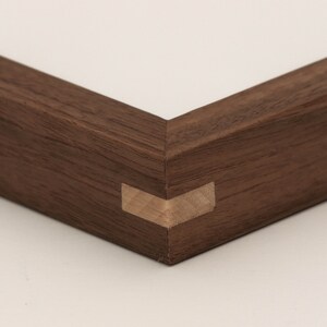 May include: Close-up of a corner of a dark brown wooden picture frame with a mitered joint. The frame is made of solid wood and has a natural finish.