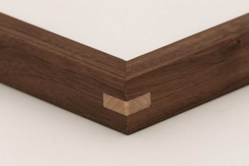 May include: Close-up of a corner of a dark brown wooden picture frame with a mitered joint. The frame is made of solid wood and has a natural finish.