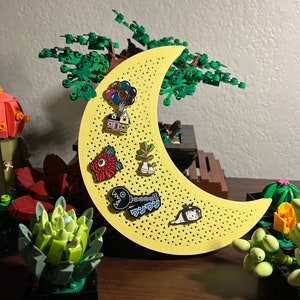 May include: A yellow crescent moon-shaped pin board with a variety of enamel pins attached. The pins include a house, a monster, a flower, a balloon, and a cat. The board is made of wood and has a textured surface.