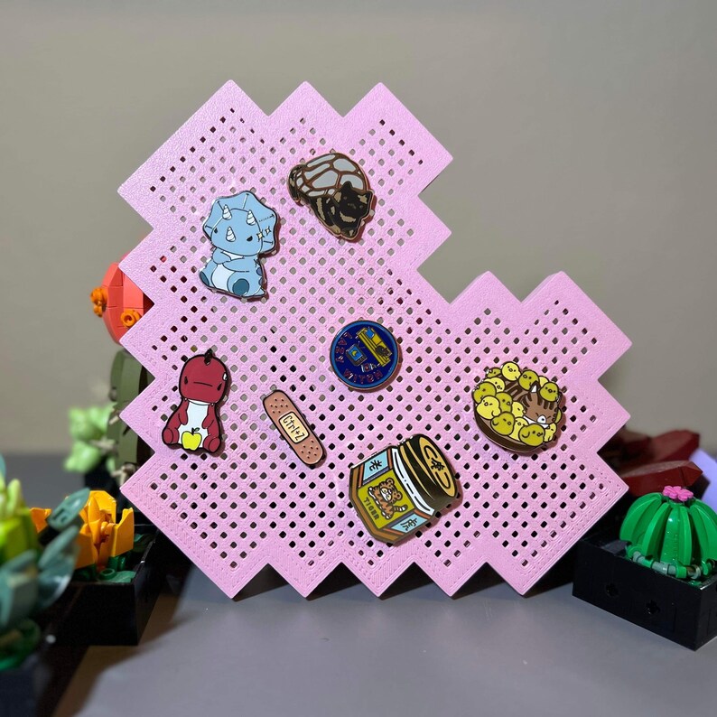 May include: A pink pegboard with a variety of enamel pins. The pins feature a dinosaur, a turtle, a plaster, a jar of honey, a group of cartoon characters, and a logo.