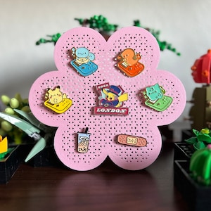 May include: A pink flower-shaped pin board with eight enamel pins. The pins feature various cartoon characters, including a Pikachu, a Squirtle, a Bulbasaur, and a Charmander. The pins are arranged in a circle around the center of the board. The board is made of a soft, pink material and has a smooth, glossy finish.