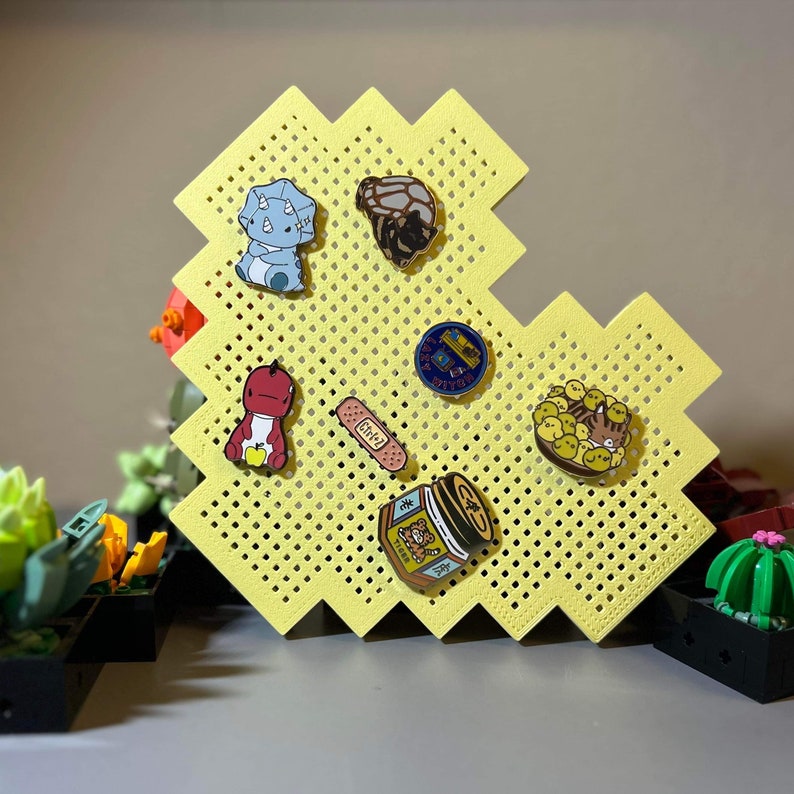 May include: A yellow pegboard with a variety of enamel pins. The pins feature a dinosaur, a turtle, a bandage, a jar of honey, a cat, and a group of chicks.