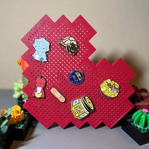 May include: A red heart-shaped pegboard with a variety of enamel pins. The pins include a dinosaur, a bandage, a jar of honey, a cat, and a group of chicks.