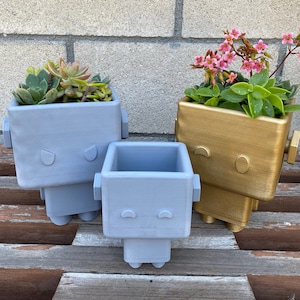 May include: Three robot-shaped planters, two are gray and one is gold. The planters are filled with succulents and flowers. The planters have a simple face design with two eyes and a mouth.