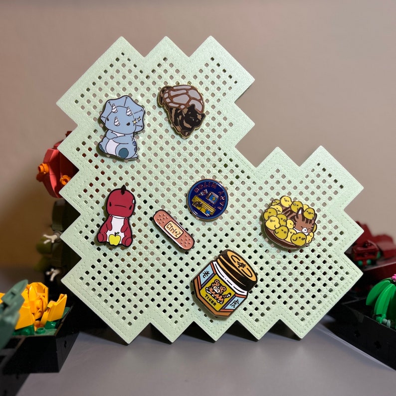 May include: A green pegboard with a variety of enamel pins. The pins feature a dinosaur, a bandage, a jar of honey, a group of chicks, a vintage arcade game, and a cartoon armadillo.