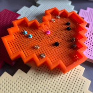 May include: Orange heart-shaped pincushion with a grid pattern. The pincushion is filled with various coloured pins, including blue, pink, gold, and black.