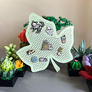 May include: A light green leaf-shaped pin board with a grid pattern.  The board is decorated with various enamel pins, including a skeleton cat, a ghost, a Totoro character, a cat with a crown, a potted plant, a boba tea, and a pin that says "cats are people".
