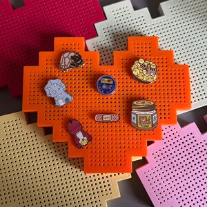 May include: An orange heart-shaped pegboard with various enamel pins. The pins feature a turtle, a cat, a jar of peanut butter, a bandage, and a cartoon character. The pegboard is on a light yellow and pink background.