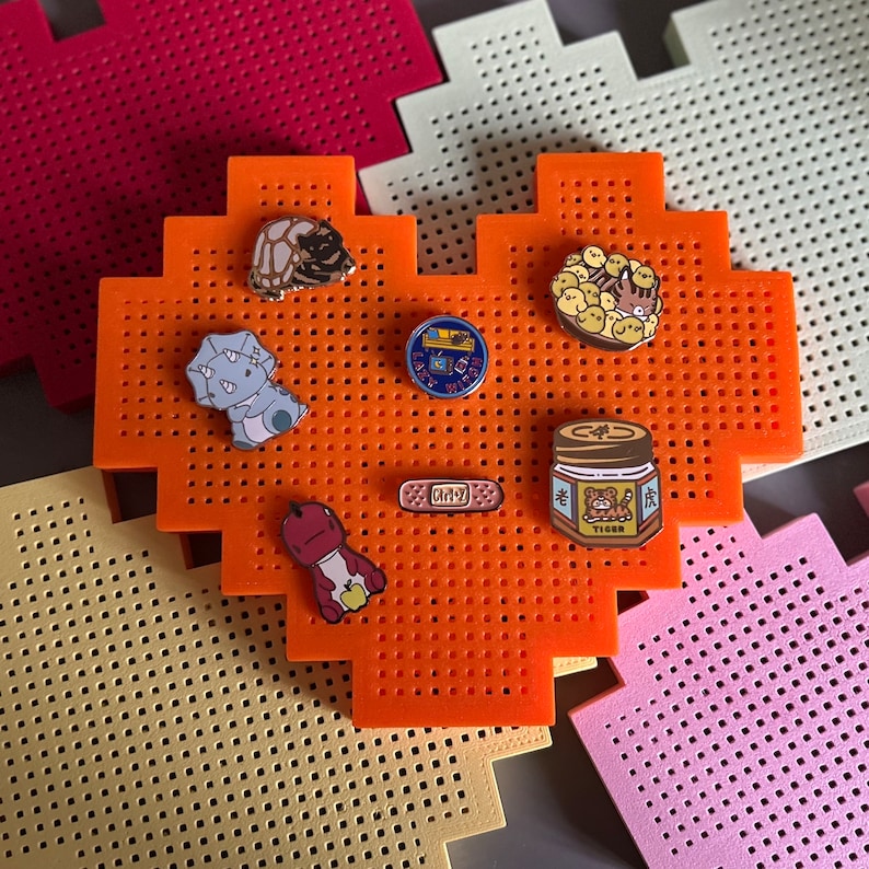 May include: An orange heart-shaped pegboard with various enamel pins. The pins feature a turtle, a cat, a jar of peanut butter, a bandage, and a cartoon character. The pegboard is on a light yellow and pink background.