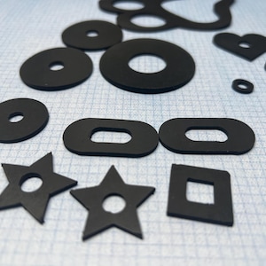 May include: A collection of black rubber washers and shapes, including circles, ovals, stars, and a heart. The various shapes have a hole in the centre. The objects are arranged on a light blue and white grid background.