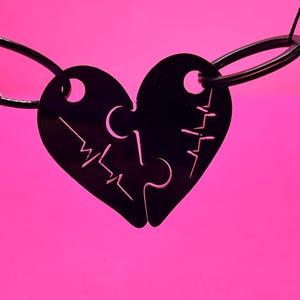 Puede incluir: Colgante negro en forma de corazón de dos piezas con un diseño de rompecabezas, suspendido sobre un fondo rosa vibrante. Cada pieza tiene un pequeño agujero para la fijación. El colgante presenta un diseño similar a un latido del corazón.