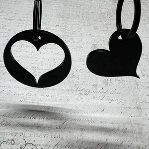 Puede incluir: Dos llaveros negros. Uno es un círculo con un corazón recortado, el otro es una forma de corazón sólido. Ambos tienen un anillo negro para sujetar a las llaves. El fondo es un papel descolorido de estilo vintage.