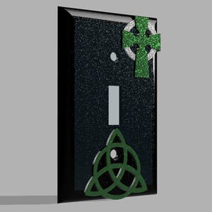 Könnte beinhalten: Eine schwarze Lichtschalterabdeckung mit einer strukturierten, glitzernden Oberfläche. Sie zeigt ein grünes keltisches Kreuz und ein grünes Triquetra-Knoten-Design. Der Schalter selbst ist ein vertikales Rechteck.