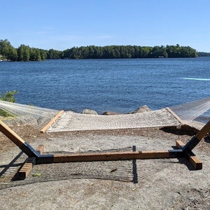 May include: A white rope hammock with a wooden frame is set up near a lake. The hammock is supported by a brown wooden frame with black metal supports. The background features a blue lake and a clear sky.