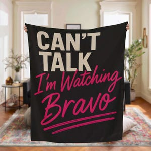 May include: Black blanket with the text "CAN'T TALK I'm Watching Bravo" in white and pink lettering. The blanket has a pink underline beneath the word "Bravo". The blanket is held up in front of a living room.