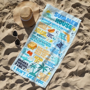 May include: A light blue beach towel with various phrases and graphics, including "Summer House," a hot dog, a sandwich, and palm trees. A straw hat and sunglasses rest on the sand next to the towel.
