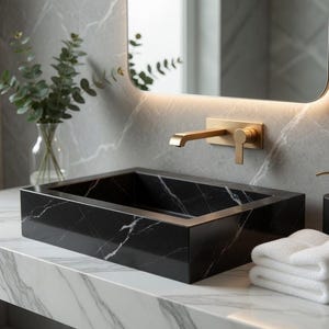 Puede incluir: Un lavabo rectangular de mármol negro y dorado en un baño moderno. El lavabo está sobre una encimera de mármol blanco, con una pila de toallas blancas y un dispensador de jabón negro. Un jarrón con vegetación y un espejo retroiluminado completan la escena.