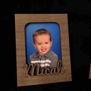 May include: A wooden photo frame with a cutout of the name Micah. The frame has a light brown stain and a photo of a young boy with a blue background.