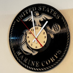 May include: A black wall clock with the words "UNITED STATES MARINE CORPS" cut out of the clock face. The clock hands are white and red, and the clock face is made of wood.