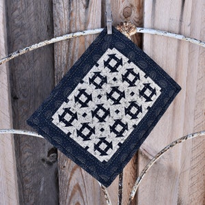 May include: A small, navy blue quilted square with a white and black geometric pattern. The square is hanging on a white metal hook.