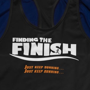 Op de afbeelding: Zwart tanktop met de tekst "FINDING THE FINISH" in wit met een hardlopend figuur en de tekst "JUST KEEP RUNNING..." in oranje. Het tanktop ligt op een blauw jack, naast een waterfles, een paar roze hardloopschoenen en een zwarte smartphone.