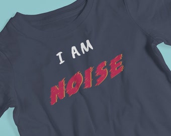 Op de afbeelding: Marineblauw T-shirt met de tekst "I AM NOISE" in wit en rood. De afbeelding toont een zonnebril en kindersandalen op een lichtblauwe ondergrond.