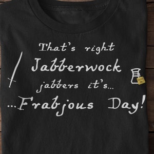 Op de afbeelding: Zwart T-shirt met witte tekst: "That's right Jabberwock jabbers it's... Frabjous Day!" Een zwaard en een beker-afbeelding zijn ook inbegrepen. Het T-shirt ligt op een houten oppervlak.
