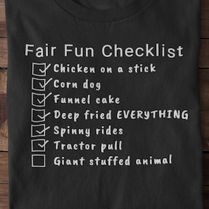 Pode incluir: Camiseta preta com uma lista de verificação branca que diz "Fair Fun Checklist" e inclui itens como "Chicken on a stick", "Corn dog", "Funnel cake", "Deep fried EVERYTHING", "Spinny rides", "Tractor pull" e "Giant stuffed animal".