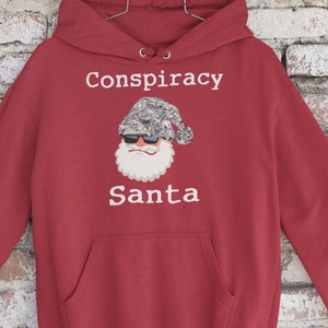 May include: Red hooded sweatshirt with a white graphic of Santa Claus wearing sunglasses and a camouflage hat. The text "Conspiracy Santa" is printed below the image.