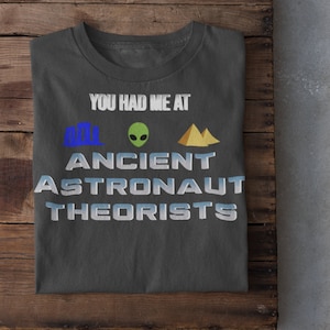 May include: A dark gray t-shirt with the text "You had me at Ancient Astronaut Theorists" in white. The text is surrounded by three icons: a blue building, a green alien, and a yellow pyramid.
