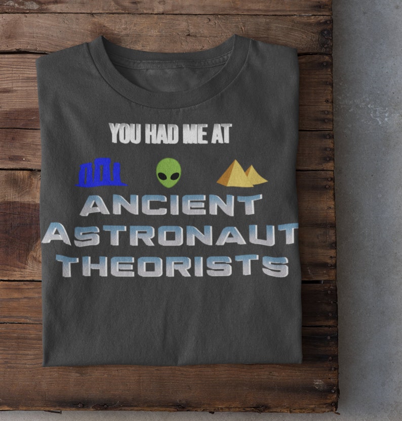 May include: A dark gray t-shirt with the text "You had me at Ancient Astronaut Theorists" in white. The text is surrounded by three icons: a blue building, a green alien, and a yellow pyramid.