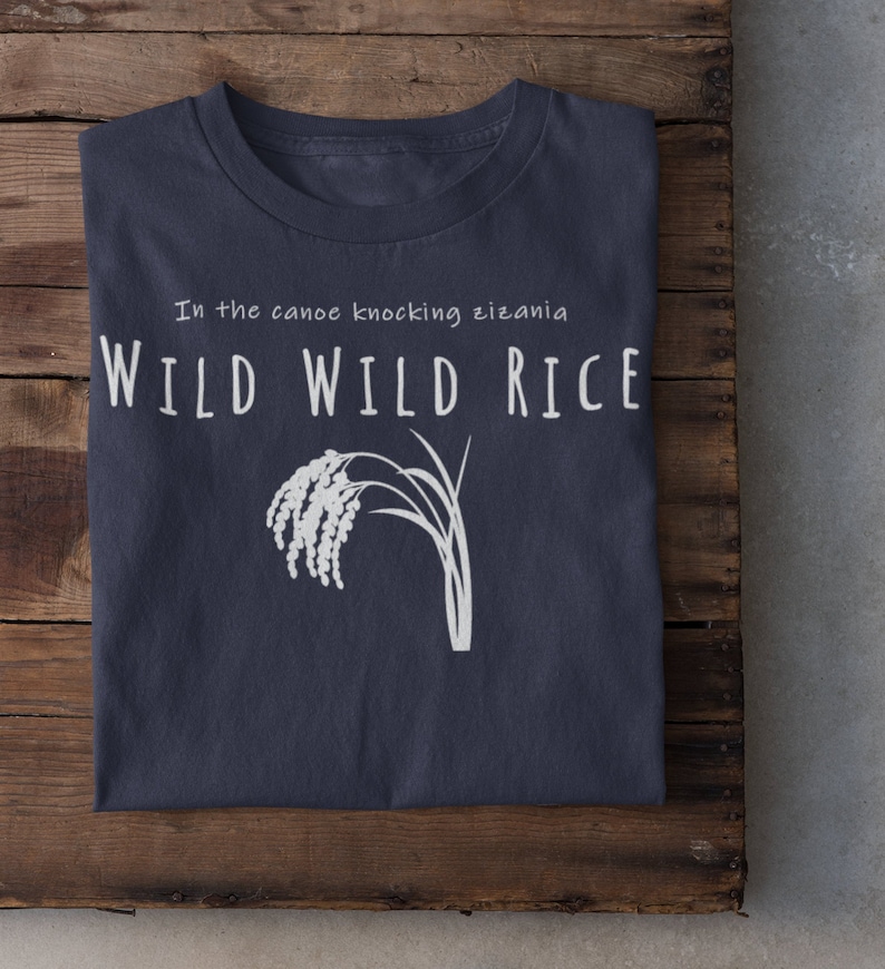 Puede incluir: Una camiseta azul marino con texto blanco que dice "In the canoe knocking zizania WILD WILD RICE" y una ilustraci&oacute;n blanca de un tallo de arroz salvaje.