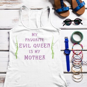 Op de afbeelding: Een wit tanktop met de tekst "My favorite evil queen is my mother" in paarse en groene letters. Het tanktop ligt op een witte houten achtergrond met blauwe sandalen, een zonnebril, een blauw horloge en armbanden.