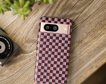Puede incluir: Una funda de teléfono con un patrón de cuadros rosas y negros. La funda está sobre una superficie de madera, junto a una cámara y una planta.