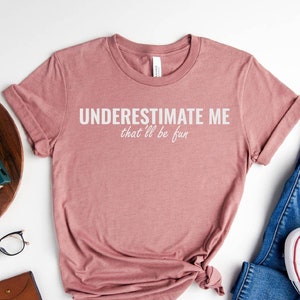 May include: A heather mauve t-shirt with the text "UNDERESTIMATE ME that'll be fun" printed in white. The shirt is shown on a white background with a pair of jeans, white sneakers, and other accessories.
