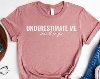 May include: A heather mauve t-shirt with the text "UNDERESTIMATE ME that'll be fun" printed in white. The shirt is shown on a white background with a pair of jeans, white sneakers, and other accessories.